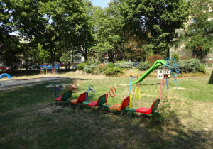 zestaw krzesełek dla dzieci z zamontowanymi kierownicami, ustawiony w rzędzie, w tle zielona zjeżdżalnia dla dzieci oraz kolorowe drabinki do wspinania i skalniak z roślnami w ogrodzie przedszkolach w